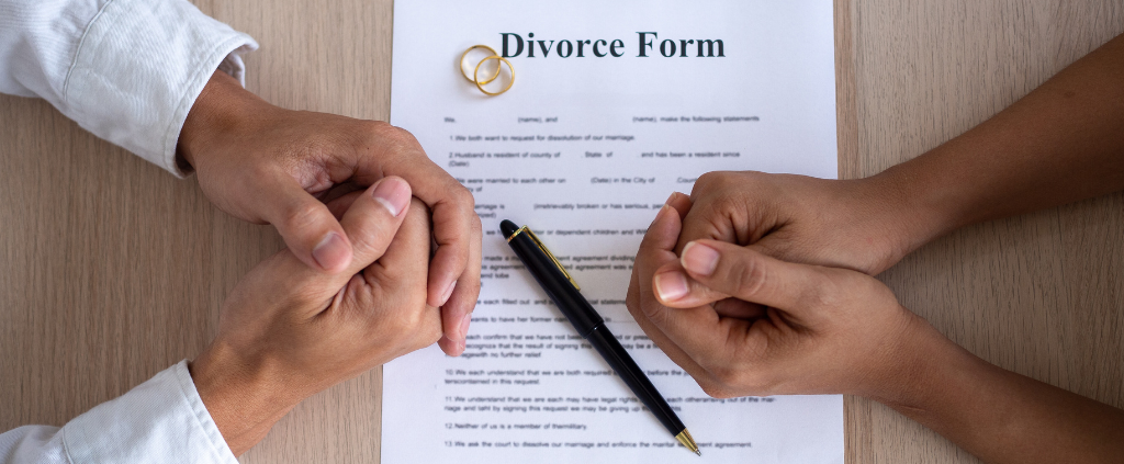 Hands clasped together over a divorce form with wedding rings and a pen.