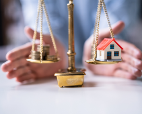 A set of gold scales balancing a miniature house on one side and a stack of coins on the other.