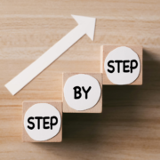 Three wooden blocks labeled “Step by Step” stacked upward with a white arrow pointing forward.