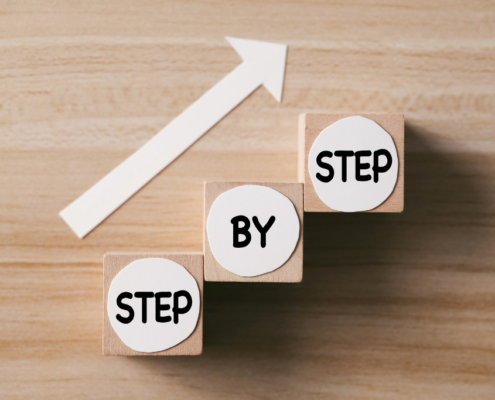 Three wooden blocks labeled “Step by Step” stacked upward with a white arrow pointing forward.