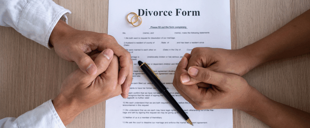 Two people’s hands clasped over a divorce form with a pen and wedding rings on a wooden table