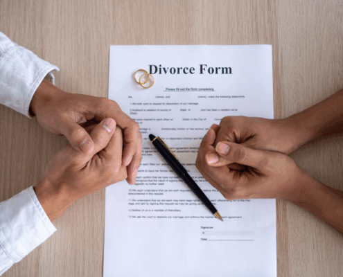 Two people’s hands clasped over a divorce form with a pen and wedding rings on a wooden table