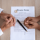 Two people’s hands clasped over a divorce form with a pen and wedding rings on a wooden table
