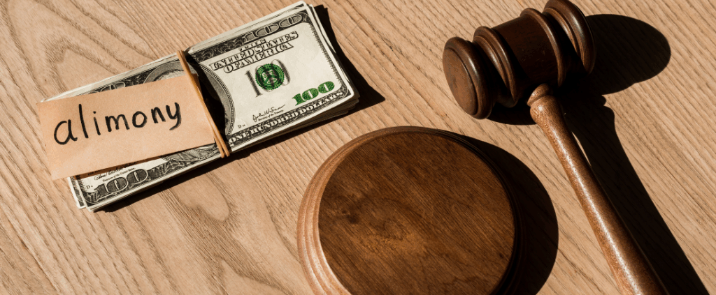 Stack of $100 bills labeled "alimony" next to judge’s gavel on wooden table.