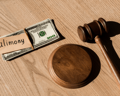 Stack of $100 bills labeled "alimony" next to judge’s gavel on wooden table.