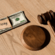 Stack of $100 bills labeled "alimony" next to judge’s gavel on wooden table.